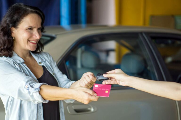 Frau bekommt Autoschlüssel gegen Bankkarte mit Auto im Hintergrund